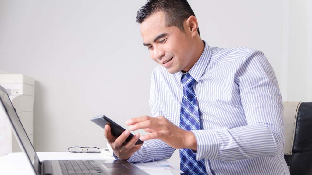 Businessperson at laptop, smiling and looking at phone